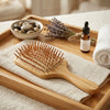 A wooden hairbrush with natural bristles rests on a folded cream towel inside a wooden tray, styled with a small amber dropper bottle, dried lavender, smooth stones, and a rolled white towel.
