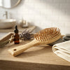 A light natural wood hairbrush with flexible bristles resting on a wooden bathroom counter, styled with a small amber dropper bottle, lavender sprigs, gray stones, and a folded cream towel in a softly blurred bathroom setting.