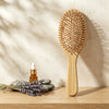 A natural bamboo hairbrush stands upright on a wooden surface against a textured beige wall with soft leaf shadows. A small amber dropper bottle, lavender sprigs, and smooth stones are arranged beside it as styled props.