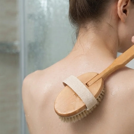 A woman with wet skin brushes her upper back with a wooden shoulder scrubber featuring a beige fabric strap.