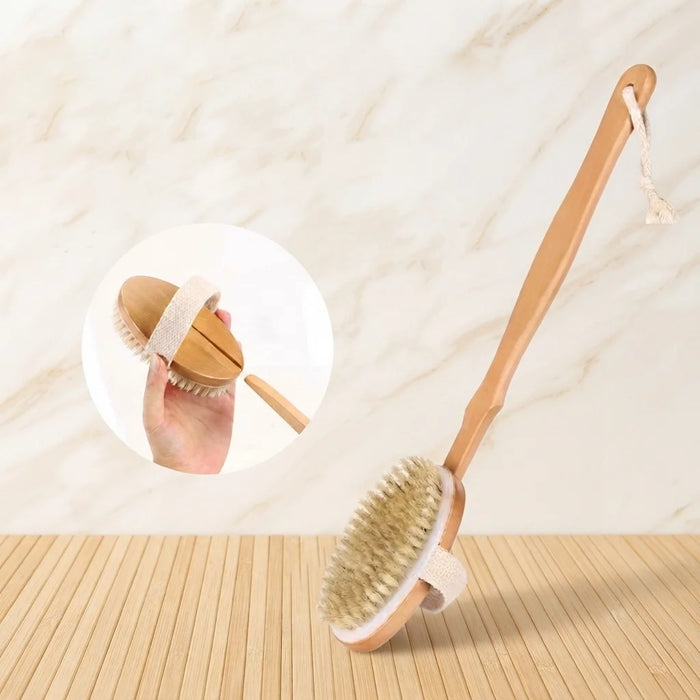 A wooden long-handled body brush with natural bristles and a fabric wrist strap, shown on a light wood surface against a marble-style background. A circular inset shows a hand holding the brush.