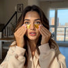 A woman in a beige fleece robe applies gold eye patches to her eyes in a brightly lit room with a window and staircase in the background.