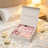 A pink silk gift set in an open white box, displayed on a bed with white roses and petals. The set includes a satin sleep mask, a matching scrunchie, and a folded fabric piece, with a warm lamp glowing in the background.
