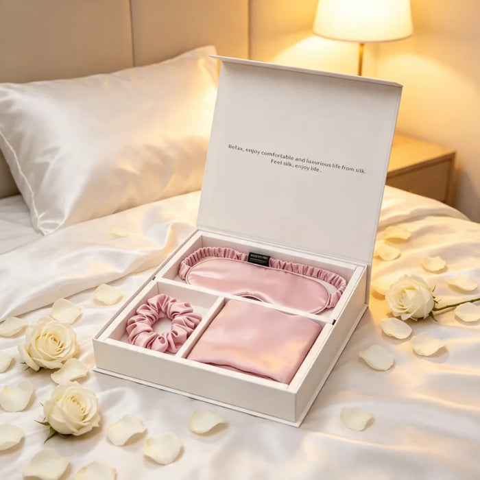 A pink silk gift set in an open white box, displayed on a bed with white roses and petals. The set includes a satin sleep mask, a matching scrunchie, and a folded fabric piece, with a warm lamp glowing in the background.