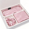 A pink silk sleep set inside a white gift box, including a satin sleep mask, a folded silk scarf, and a matching scrunchie.