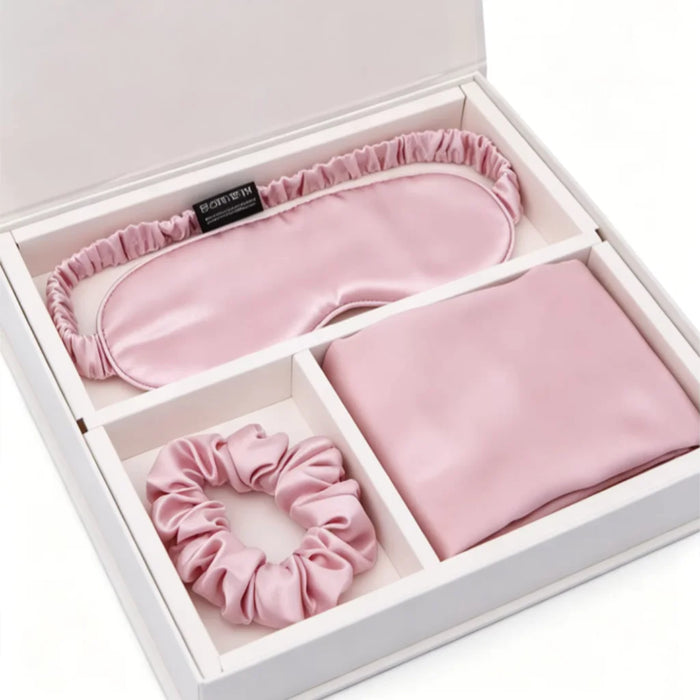 A pink silk sleep set inside a white gift box, including a satin sleep mask, a folded silk scarf, and a matching scrunchie.