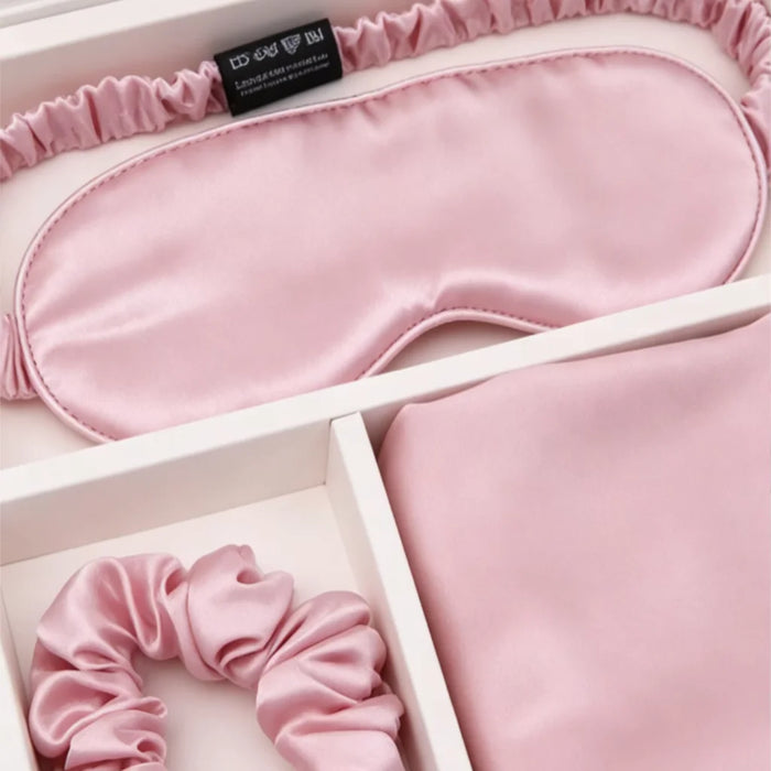 A pink silk sleep mask and matching scrunchie displayed in a white gift box with a folded pink fabric item, likely a hair tie or scarf.