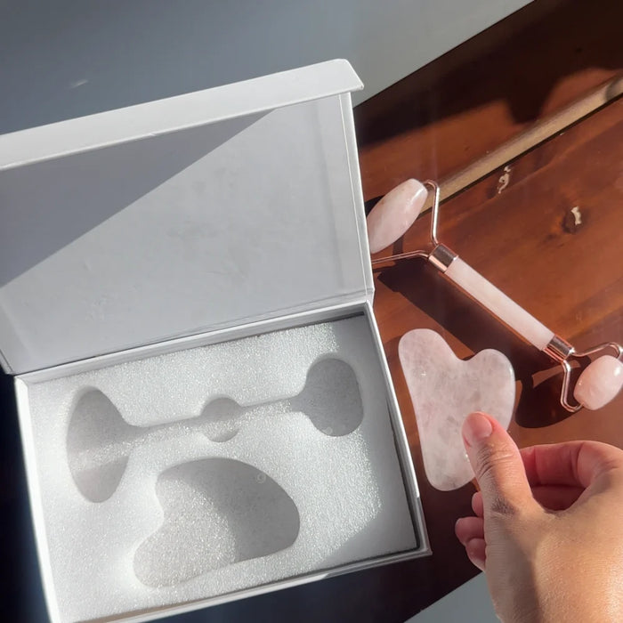 Rose quartz facial roller and gua sha set shown next to its white gift box with foam insert, held by a hand on a wooden table.