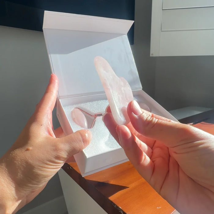A person holds a translucent pale pink rose quartz facial roller tool in front of an open white gift box containing the matching rose quartz tongue scraper, set on a wooden table.