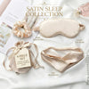 A satin sleep set arranged on white fabric with text labels identifying the components: a satin sleep mask, satin eye scrunchie, silk headband, and a small pouch with a tag reading '4-PIECE SET.' A perfume bottle and magazine are partially visible as styling props.