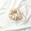 A satin hair scrunchie in a light champagne beige color, displayed on a draped white fabric background with a small section of blonde hair next to it and the text 'HAIR SCRUNCHIE' printed below.