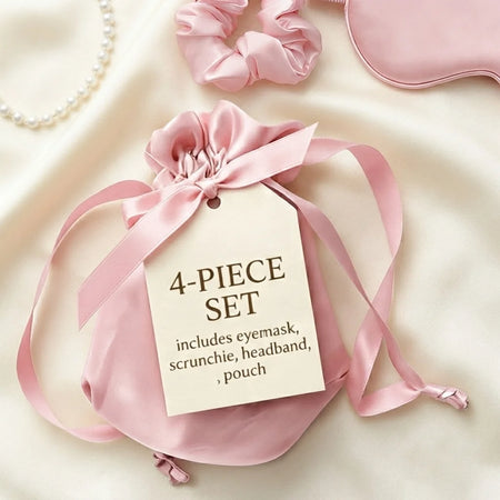 A pink satin 4-piece set gift pouch with a drawstring and ribbon, attached to a paper tag that reads '4-PIECE SET includes eyemask, scrunchie, headband, pouch.' Styled on a cream satin background with partial views of a pink eye mask, pink scrunchie, pearl necklace, and a pink headband.