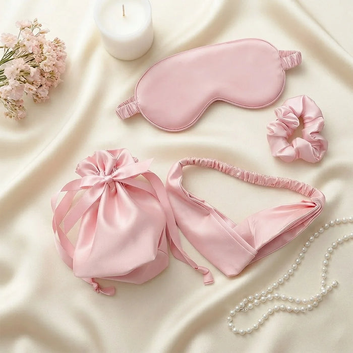 A coordinated pink satin self-care set arranged on cream satin fabric, including a sleep mask, scrunchie, drawstring pouch, headband, and pearl necklace. A white candle and small pink flowers are also present as styled props.