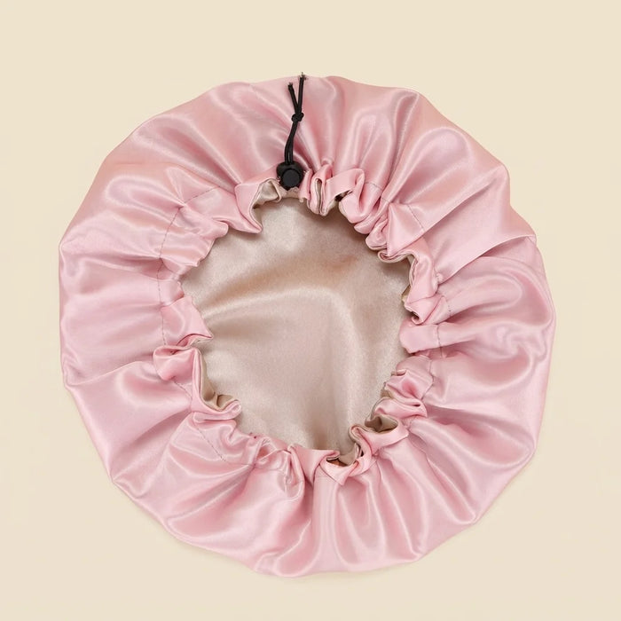 A light pink satin hair cap with a drawstring closure, laid flat on a solid beige background.