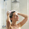 A woman washing her hair under a shower head in a glass shower stall, using a pink and white hair brush with soap lather.
