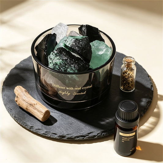 The ZenPulse Luminous Crystal Diffuser Hilton Garden variant, featuring a dark glass bowl filled with raw green and black crystals. The aromatherapy set is presented on a black slate tray with a small vial of dried botanicals, a piece of natural wood, and an essential oil bottle under soft natural shadows.
