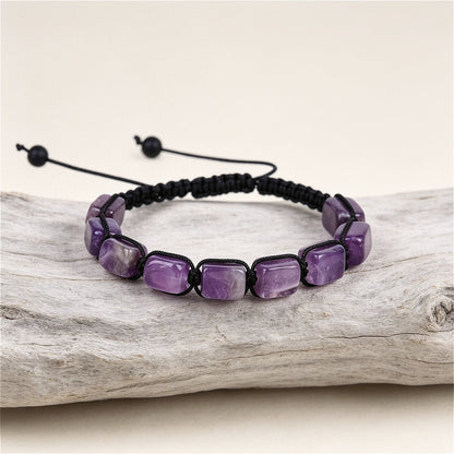 A hand-woven adjustable bracelet featuring polished rectangular purple amethyst gemstone pillars, from the ZenPulse Element Collection, model Deep Serenity. The bracelet is displayed on a rustic light wood branch against a soft beige background.
