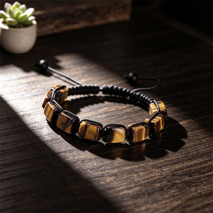 A hand-woven adjustable bracelet featuring polished rectangular Tiger Eye gemstone pillars from the ZenPulse Element Collection. The Focus & Willpower model is showcased on a dark wooden surface under dramatic natural lighting, highlighting the golden-brown chatoyancy and unique banded textures of the stones.