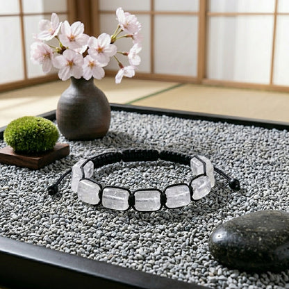 A hand-woven adjustable bracelet featuring polished rectangular White Crystal gemstone pillars from the ZenPulse Element Collection. The Pure Clarity model is elegantly placed on a bed of fine grey pebbles in a Japanese-inspired Zen garden setting, with a cherry blossom vase and shoji screen background, showcasing the icy transparency of the stones.