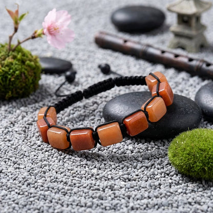 A hand-woven adjustable bracelet featuring polished rectangular Red Aventurine gemstone pillars from the ZenPulse Element Collection. The Vitality Drive model is showcased in a peaceful Zen setting on smooth black river stones and grey pebbles, accompanied by a cherry blossom and a traditional flute, highlighting the warm, sunset-orange tones of the minerals.